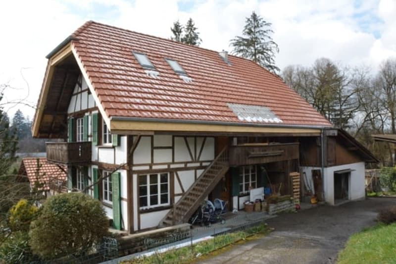 Two-story house with red-tiled roof, white and green trim, multiple windows, and a wooden staircase leading to a balcony. The house has a mix of traditional and modern architectural elements, with a small attached garage and outdoor storage area.