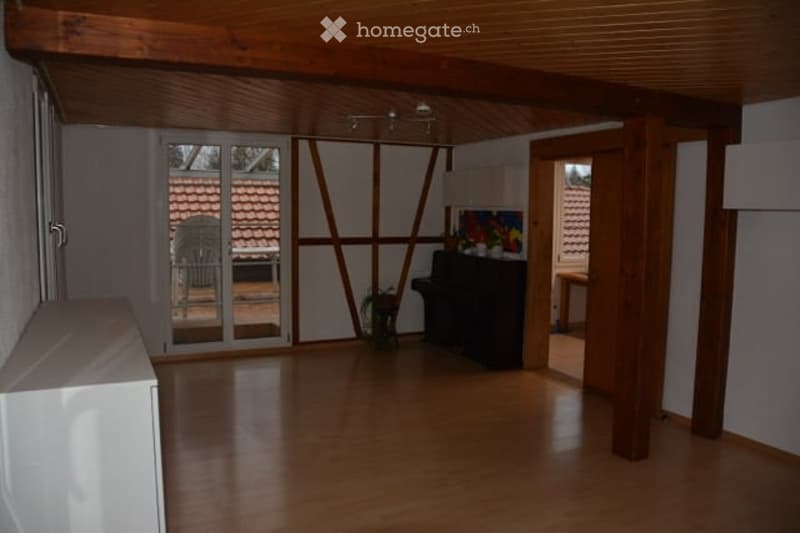 Bright, open room with wooden floors and exposed wooden beams. Features a grand piano, a small table, a chair, and a potted plant near the large glass door leading to a patio.