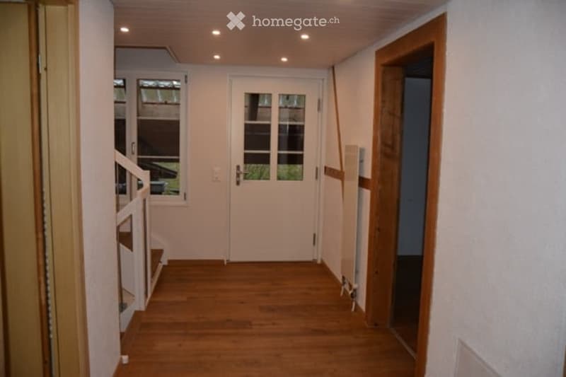 A narrow hallway with wooden flooring, white walls, and recessed ceiling lighting. Features a white front door with glass panels and a staircase leading upwards.