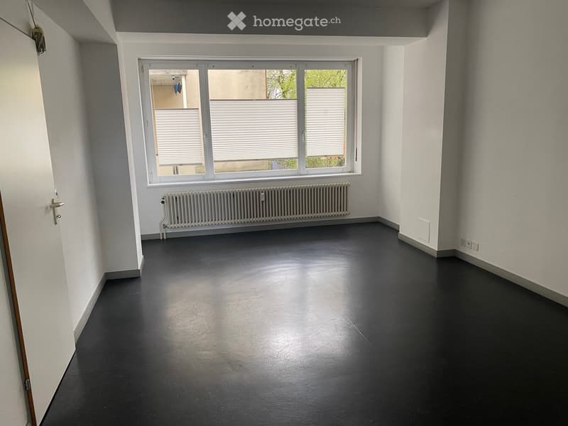 Empty room with white walls, dark floor, large window with blinds, radiator, and a door.