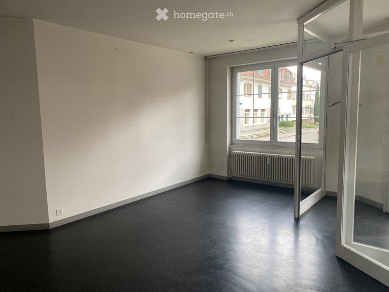 Empty room with white walls, dark flooring, large window with view of street, radiator, and glass door.