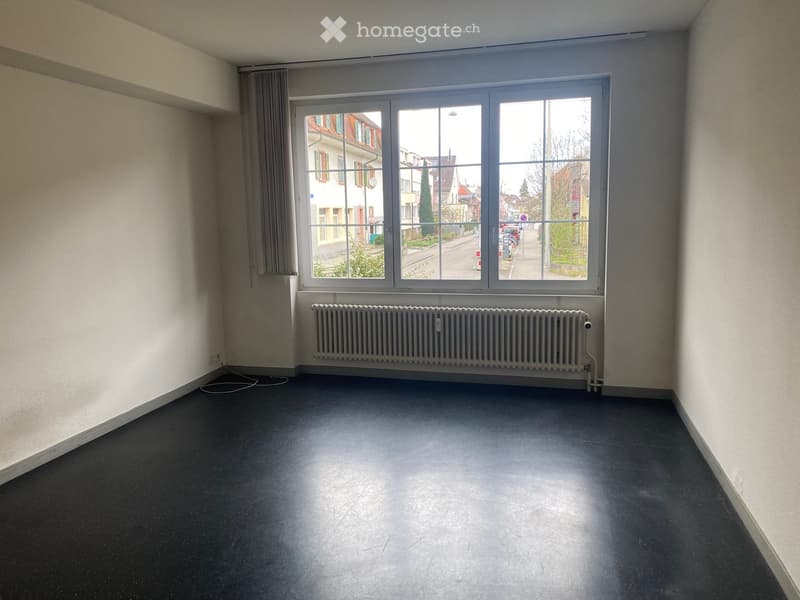 Empty room with dark flooring, white walls, large window with view of street, radiator below window, and a curtain on one side.