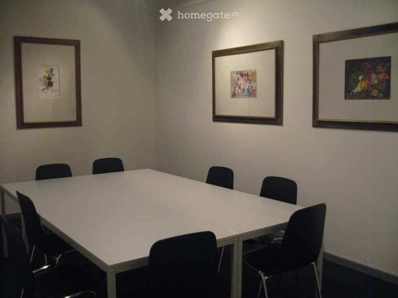 A simple, unfurnished meeting room with a large white table and several black chairs, three framed artworks on the wall.