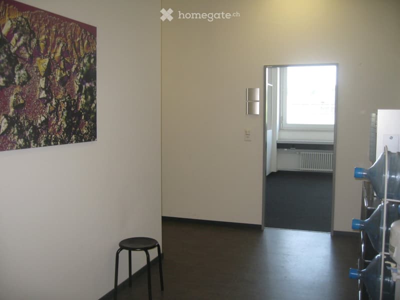 Empty room with light-colored walls, dark flooring, and a large abstract painting on one wall. A small black stool is near the entrance, and a water cooler is visible in the adjacent room.