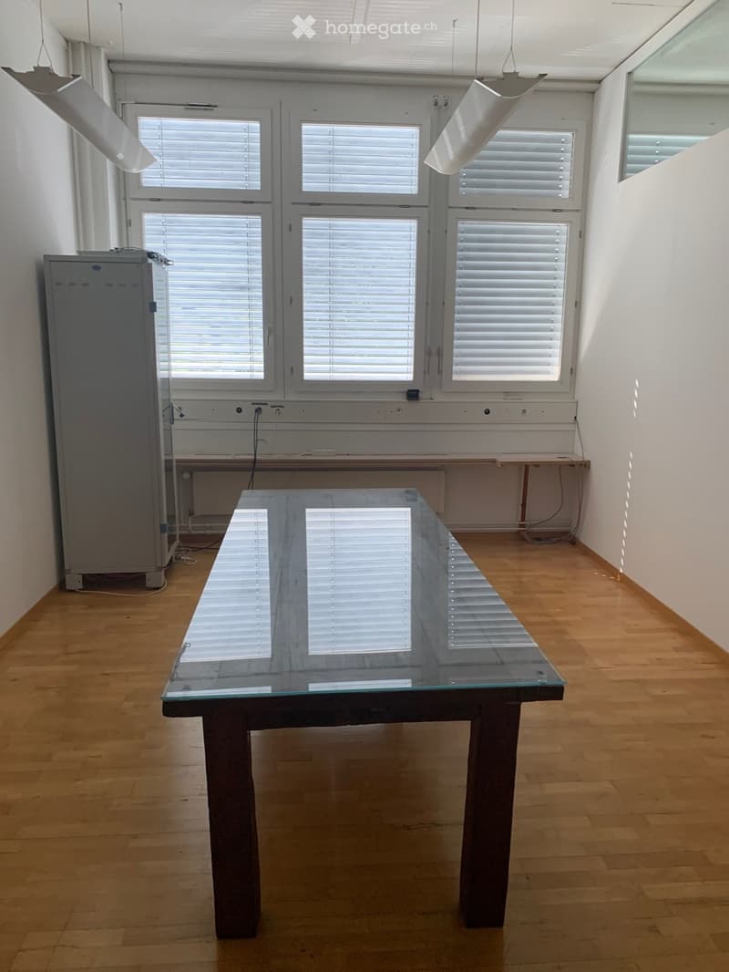 Bright, minimalist room with wooden flooring, large windows with blinds, a glass-top table, and a refrigerator.