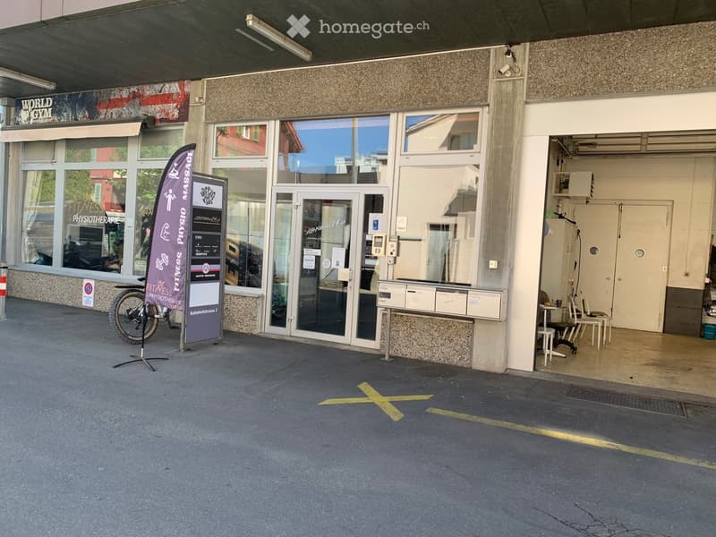 Building entrance with glass doors, signage for "Fitness & Physiotherapie," a banner, and a small seating area inside.