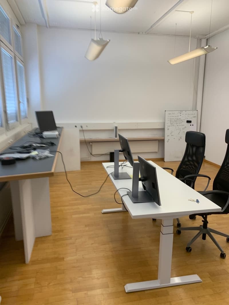 Modern office space with wooden flooring, white walls, and large windows. Includes desks, chairs, laptops, and a whiteboard.