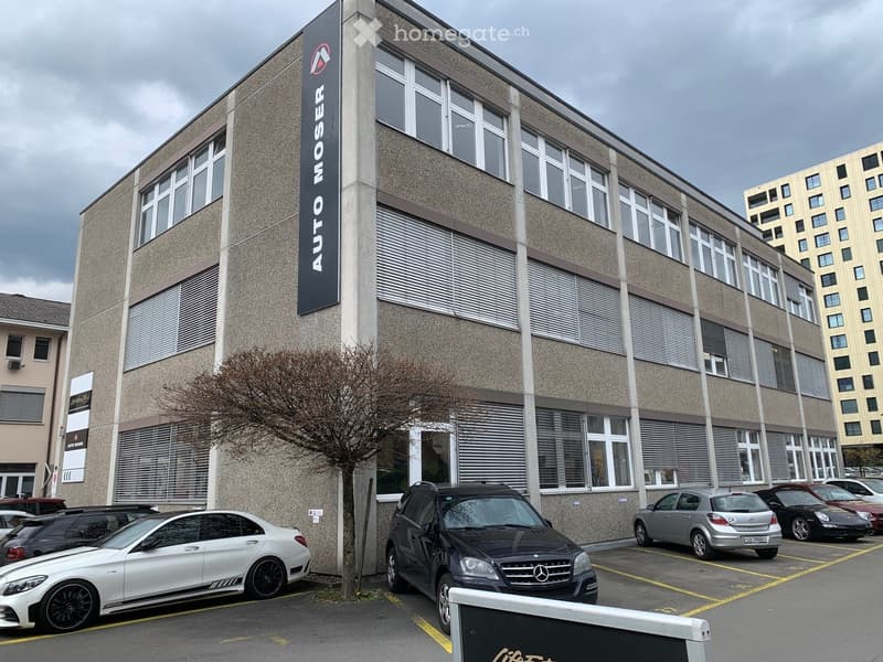 Multi-story building with large windows and a sign reading "AUTO MOSER" on the facade, with several cars parked in front.