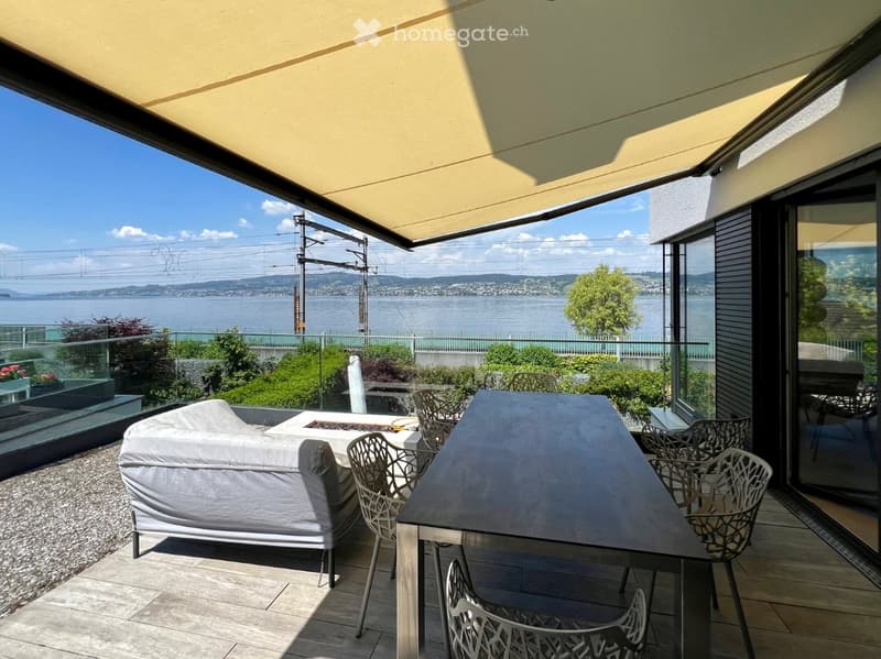 Balcony with a covered area, sofa, dining table, chairs, and a scenic view of a lake and mountains.