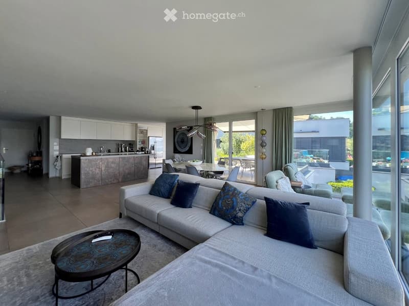 Modern living room with a grey sectional sofa, coffee table, and a kitchen area with a kitchen island, chairs, and a dining table. Large windows offer a view of the outdoor area.