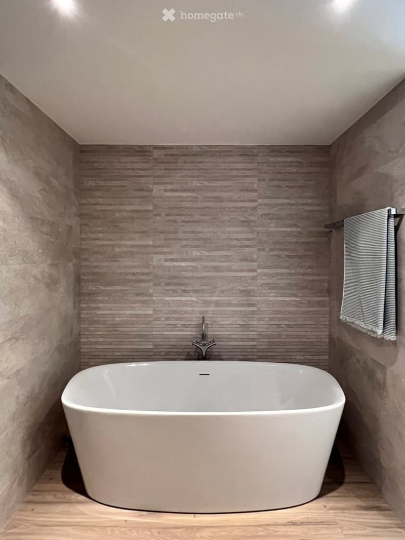 Modern bathroom with a freestanding bathtub, sleek faucet, and a towel hanging on a rack. The walls are tiled in a wood-like pattern, and the floor is wooden.