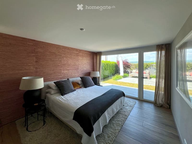 Modern bedroom with a large bed, nightstands, lamps, and large windows offering a view of the garden and sea.