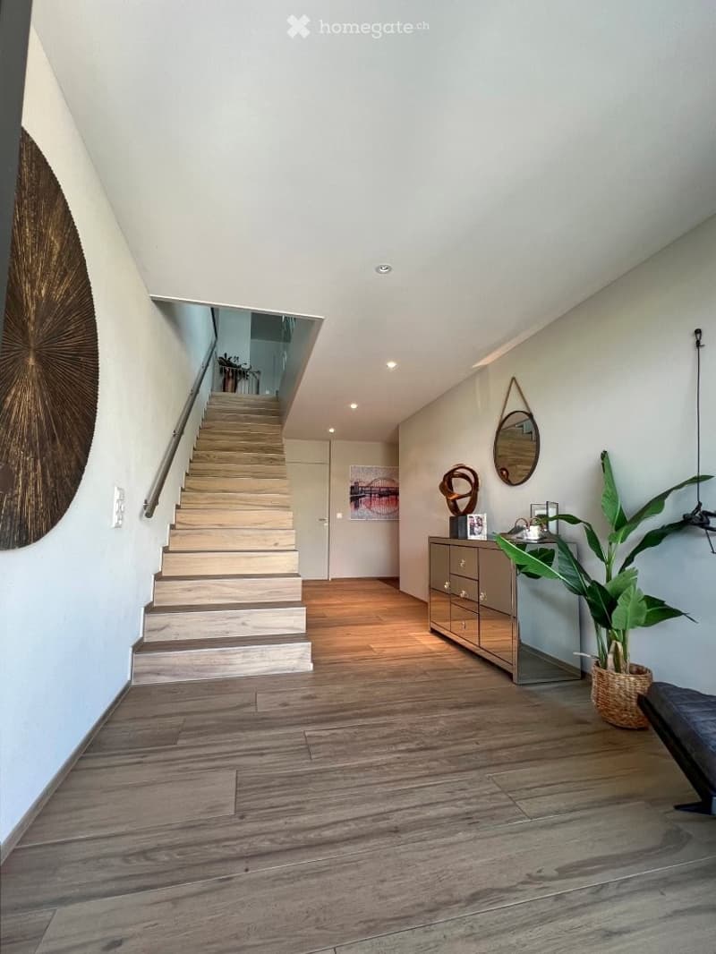 Modern hallway with wooden flooring, a staircase leading upwards, a small console table with a plant, and decorative wall art.