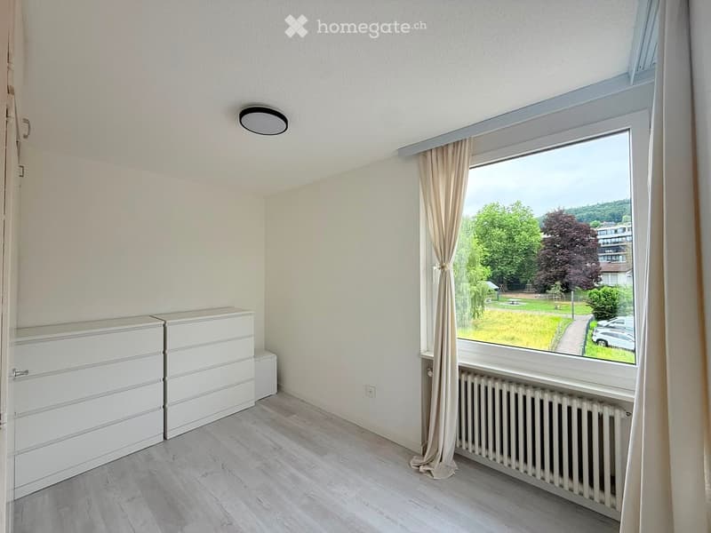 Empty room with light-colored walls, wooden flooring, a large window with curtains, a radiator, and a built-in wardrobe unit.