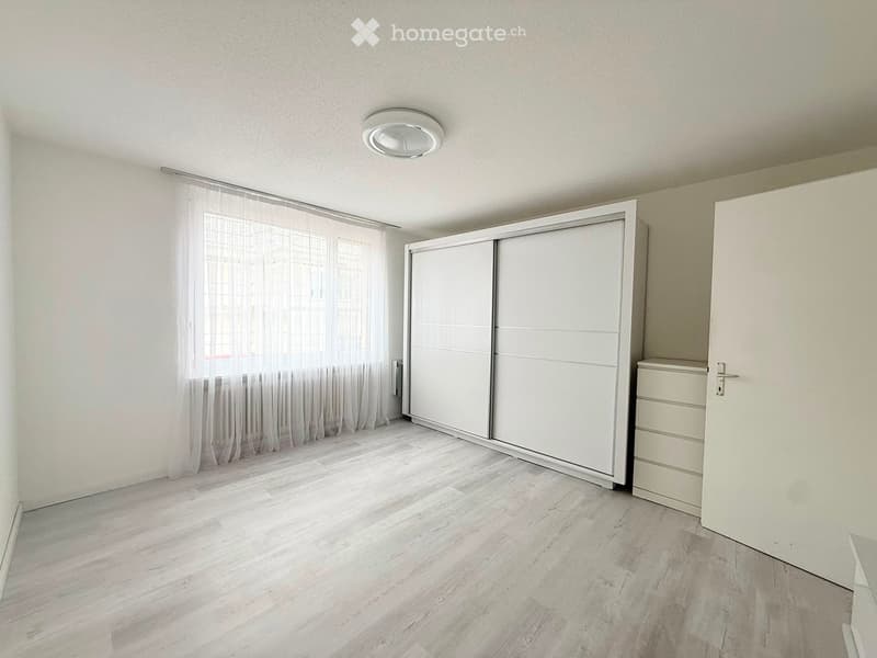 Empty room with light wooden flooring, white walls, large window with sheer curtains, built-in wardrobe, and a small chest of drawers.