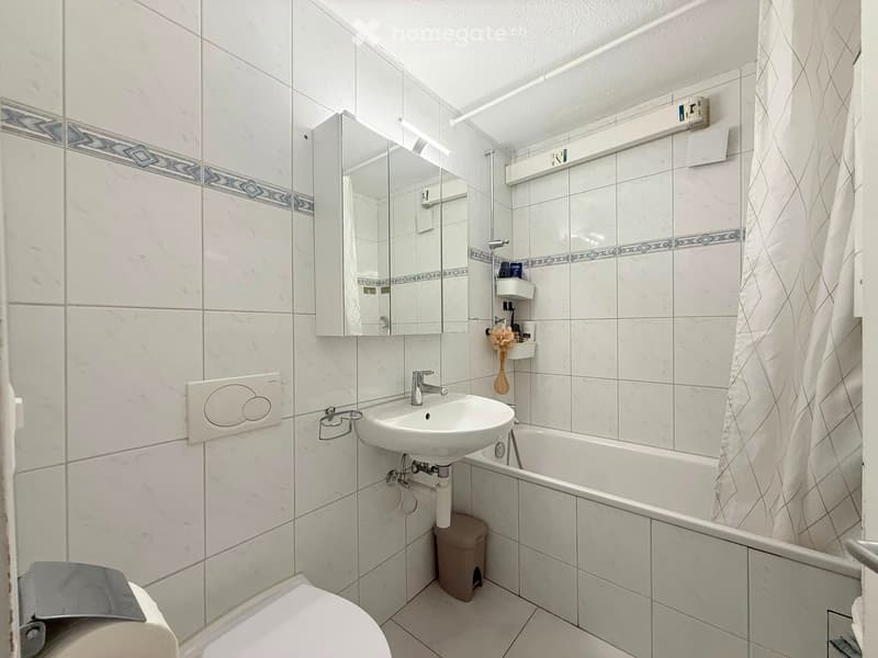 Modern bathroom with white tiles, a sink, bathtub, toilet, mirror, and a shower curtain. Includes a towel, toilet paper holder, and a small shower shelf with toiletries.