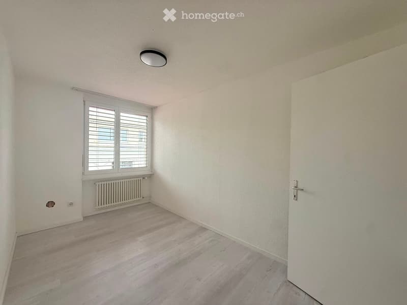 Empty room with light-colored walls, wooden flooring, a window with blinds, a radiator, and a closed door.