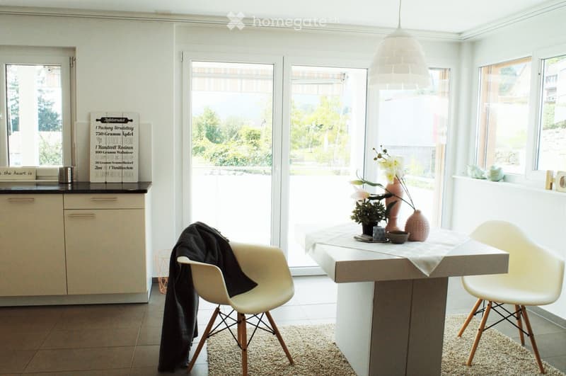 Modern kitchen with white cabinets, a central island table, two white chairs, decorative vases, a bowl, a candle, and a sign on the counter. Large windows and a sliding glass door lead to a view of greenery outside.