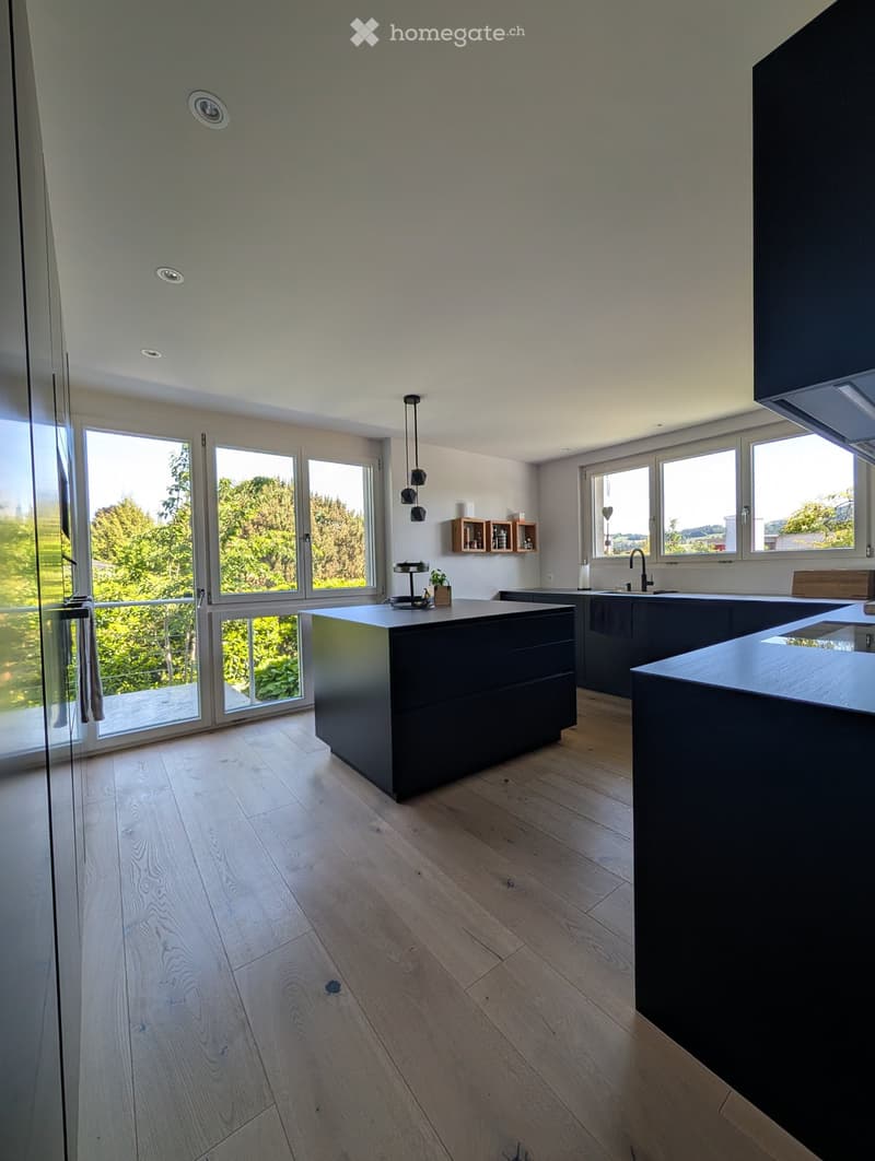 Modern kitchen with light wooden flooring, dark island, and large windows offering a scenic view.