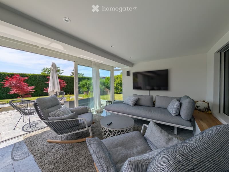Modern living room with large windows, grey sofa, coffee table, chairs, and television.