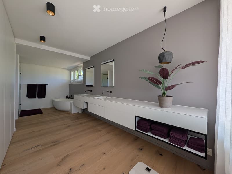 Modern bathroom with a white vanity, double sinks, a freestanding bathtub, and a potted plant on the counter. The room features wooden flooring, grey walls, and built-in storage shelves with neatly folded towels.