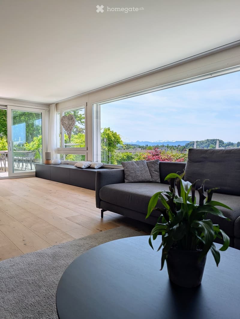 Modern living room with large windows offering a scenic view, featuring a grey sofa, coffee table, potted plant, and side table.