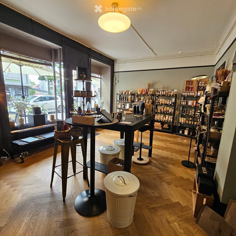A well-lit, modern retail or cafe space with wooden flooring, large windows, shelves filled with products, a central table with a laptop, and decorative items.