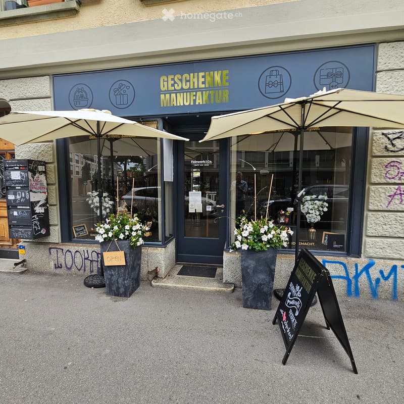 Storefront with a sign reading "GESCHENKE MANUFAKTUR" and two large umbrellas over the entrance. Potted plants and a chalkboard sign are visible.