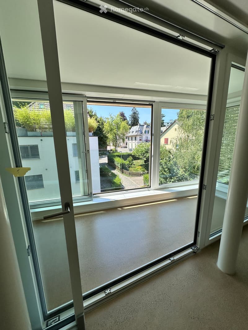 Empty room with large windows and a view of the street and surrounding greenery. The room has a smooth, light-colored floor and white walls.