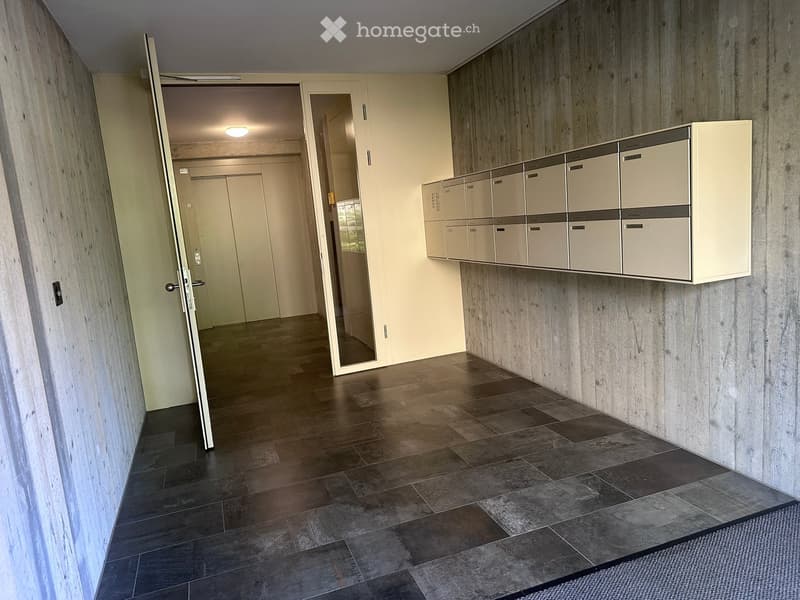 Modern hallway with wooden and tiled flooring, white walls, and a row of mailboxes mounted on the wall. An open door leads to another area.