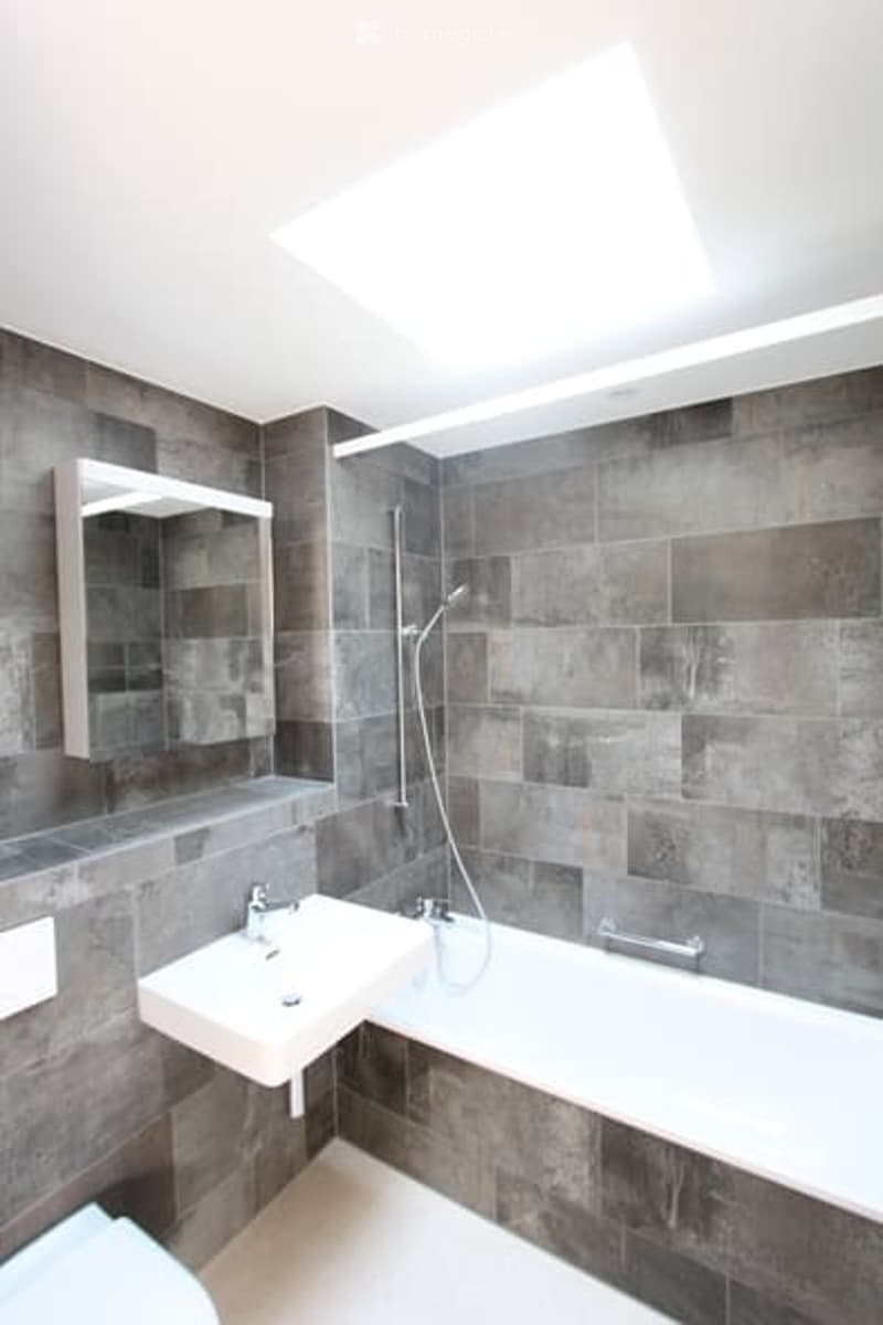A grey tiled bathroom with a white bathtub, a white vanity, a mirror, and a white toilet.
