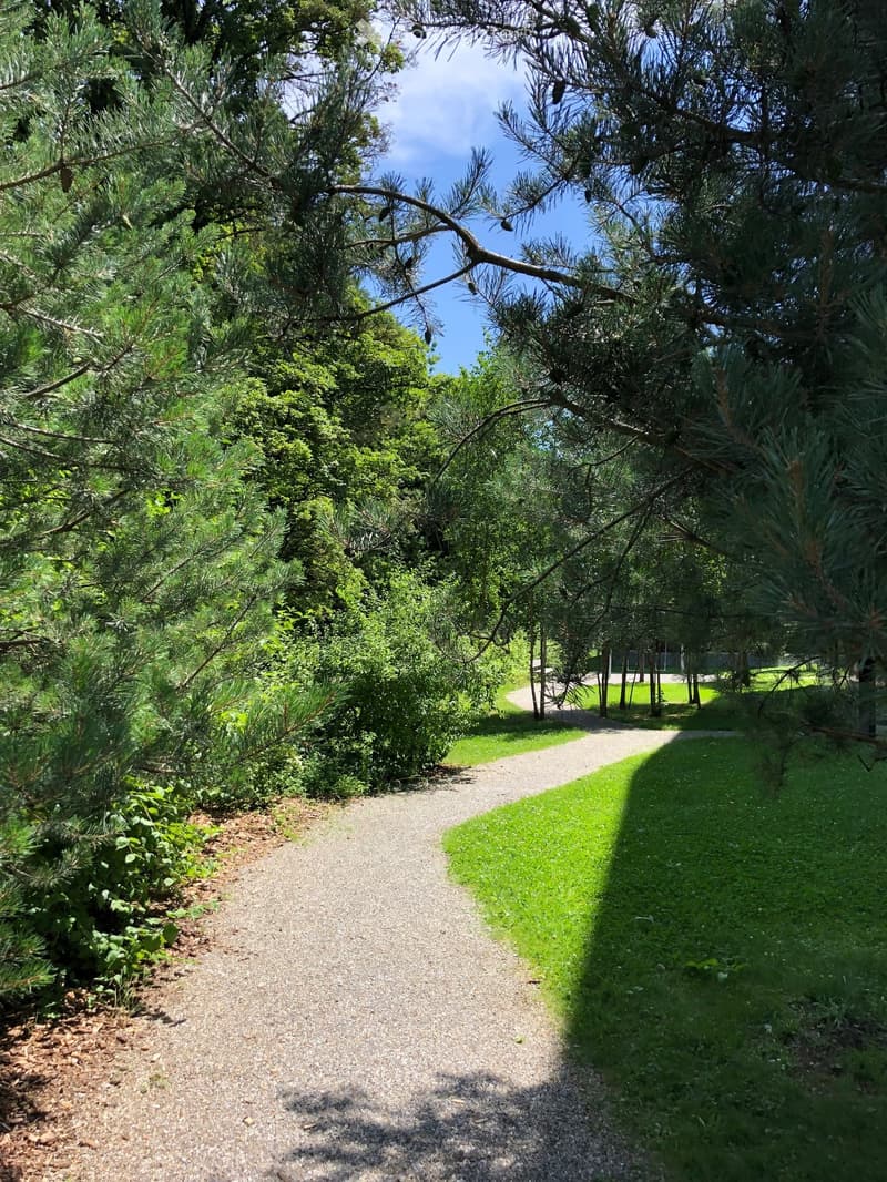 gravel pathway through a tree lined garden, surrounded by grass, in the distance, a tennis court.