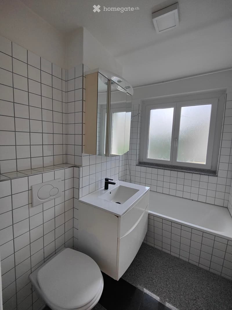 Modern bathroom with white tiled walls, a white sink, a bathtub, a toilet, and a large mirror above the sink. The room features a window with frosted glass and a sleek, contemporary design.