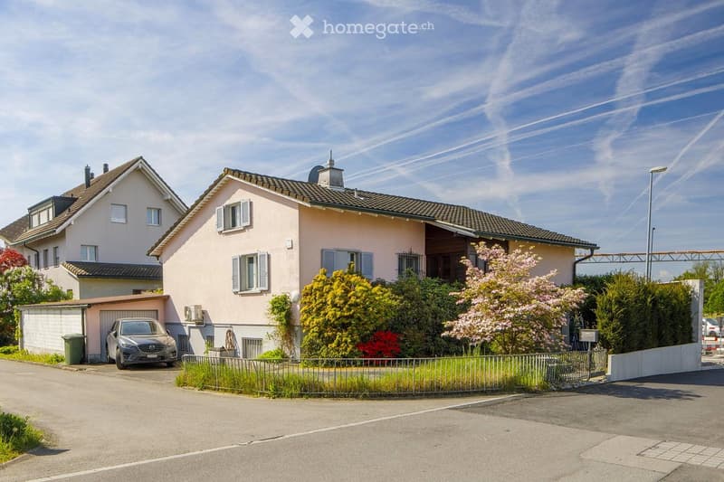 Two-story house with beige exterior, multiple windows, garage, and well-maintained garden with flowering trees and shrubs.