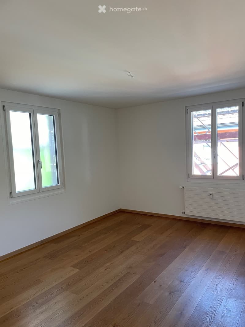 Empty room with wooden flooring, white walls, and two windows with white frames. A white radiator is visible on one wall.