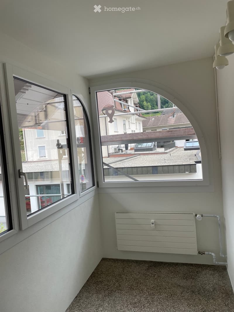 Empty room with light-colored walls, a radiator, and large windows offering views of neighboring buildings and a balcony area.