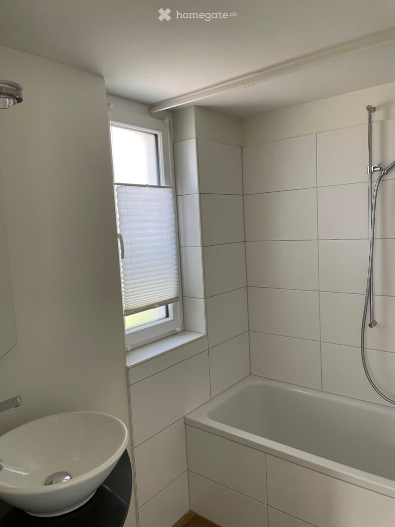 Modern bathroom with a bathtub, sink, and showerhead. Light-colored tiles and a window with blinds provide natural light.