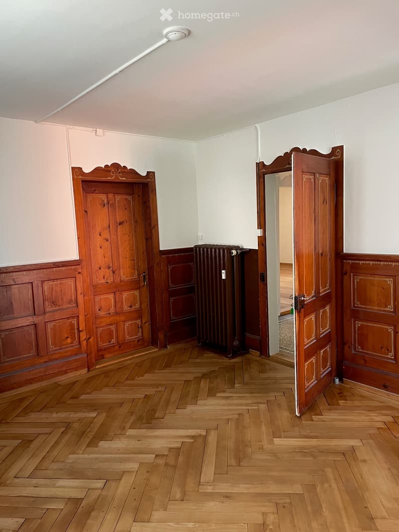 Empty room with wooden herringbone flooring, white walls, and two wooden doors with ornate carvings. A radiator is visible against one wall.