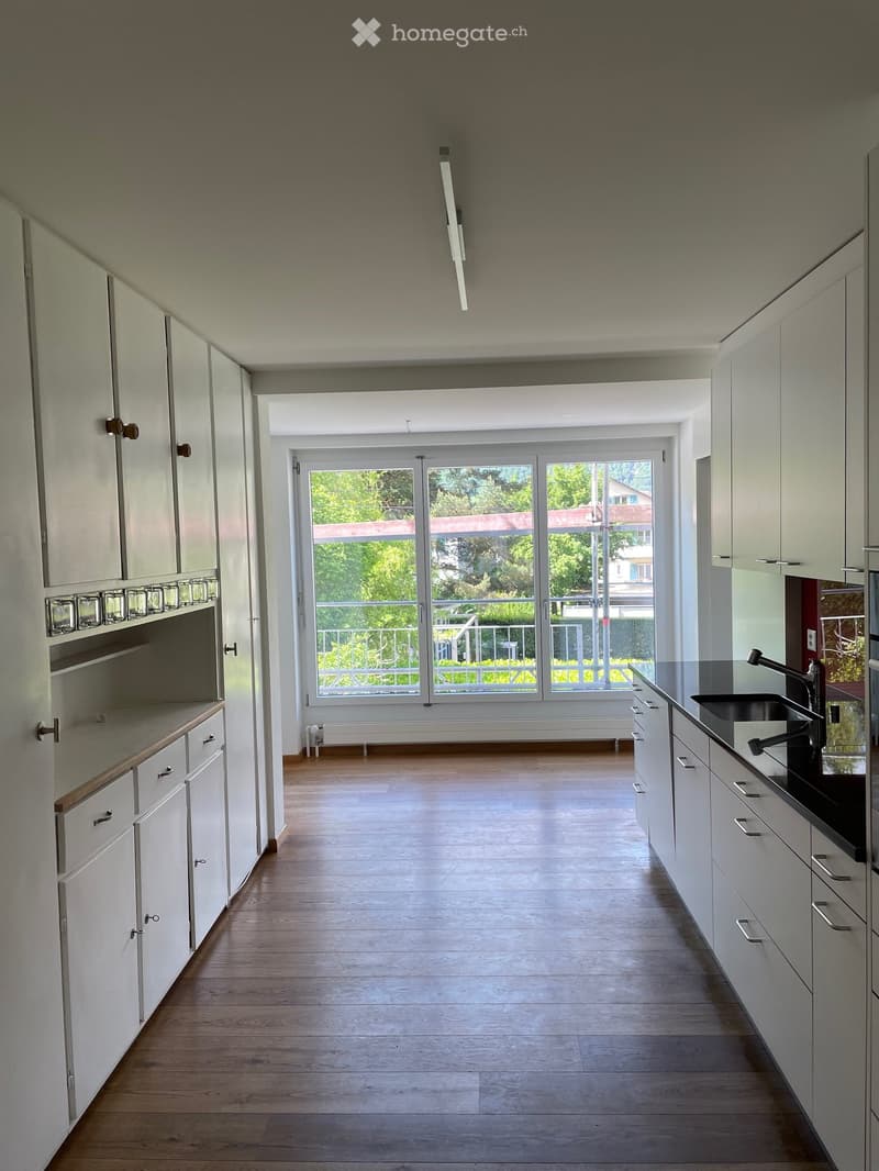 Modern kitchen with white cabinets, wooden flooring, and a large window leading to a balcony.