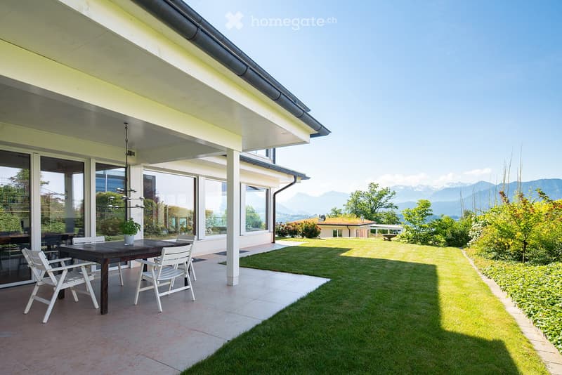 Outdoor patio area with a wooden table, white chairs, and a potted plant, overlooking a well-maintained lawn and distant mountain view.