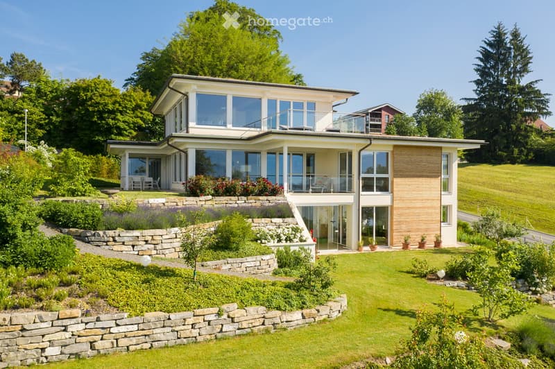 Modern two-story house with large windows, balconies, and a well-maintained garden with stone walls and greenery.