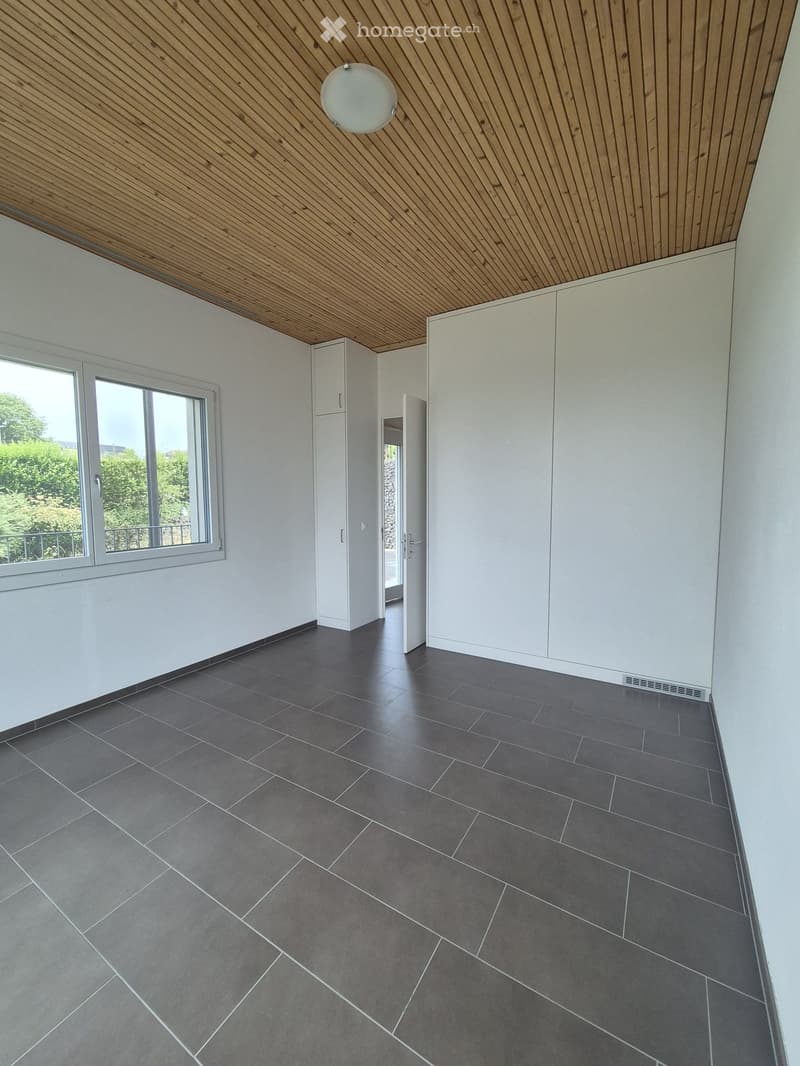Empty room with dark grey tiled floor, white walls, large window with view of greenery, white built-in cabinets, and a modern pendant light fixture on a wooden slanted ceiling.