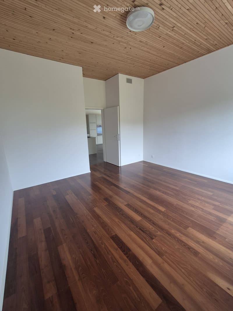 Empty room with wooden flooring, white walls, and a wooden ceiling. A doorway leads to another room with white cabinets and a built-in oven.