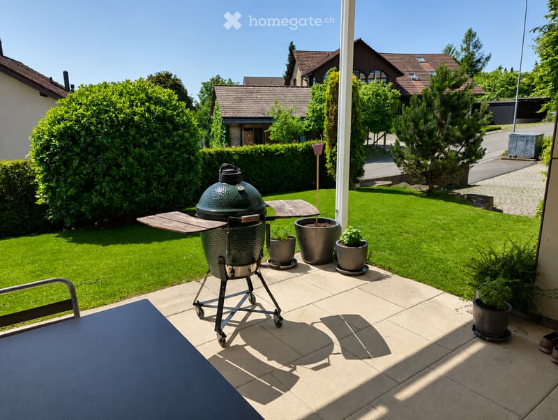 Well-maintained garden with a large green barbecue grill, potted plants, and neatly trimmed hedges. The patio features a blue table and chairs, with a clear view of neighboring houses and a paved driveway.