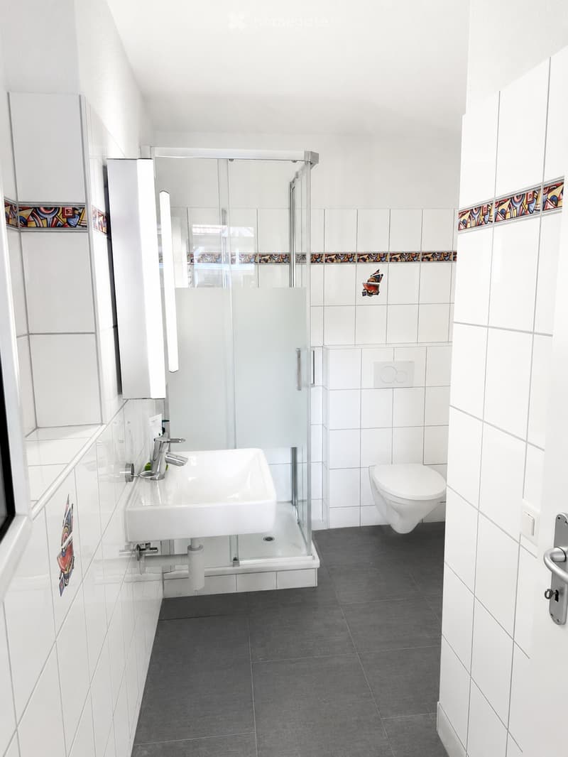 Modern bathroom with white tiles, a glass-enclosed shower, a white sink with a chrome faucet, and a toilet. The walls feature decorative tiles with colorful patterns.
