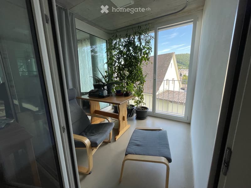 Balcony with a wooden table, two chairs, a stool, and several potted plants. Large windows offer a view of the surrounding area.