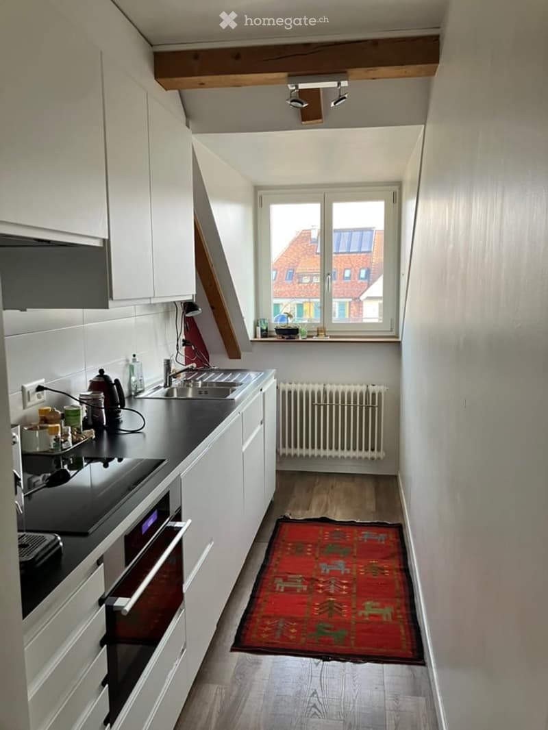 modern attic kitchen with wooden floor, white cabinets, black countertop, large window, sink with faucet, radiator, rug, modern appliances