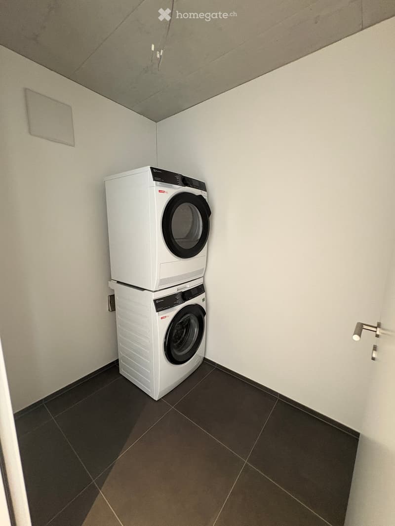 A small, clean laundry area with a stacked washing machine and dryer, dark tiled floor, and white walls.