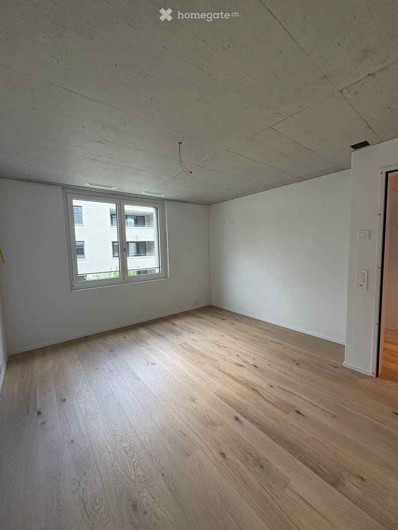 Empty room with light wooden flooring and white walls, featuring a large window with a view of neighboring buildings.