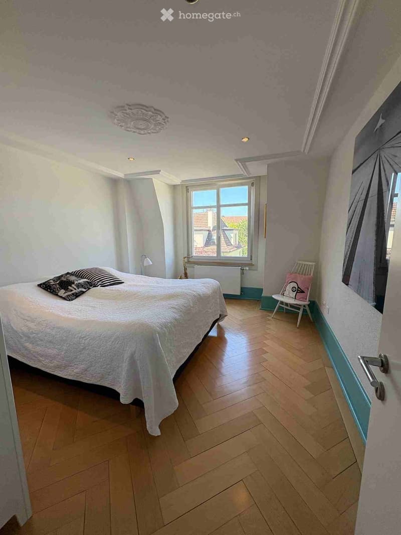 Cozy bedroom with wooden flooring, a large bed with white bedding and black and white striped pillows, a small white chair with a pink cushion, and a window with white curtains. The walls are painted in soft pastel colors, and the room features a decorati